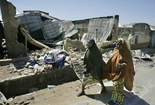 Nigerian displaced women walk past a destroyed house {PressTv}