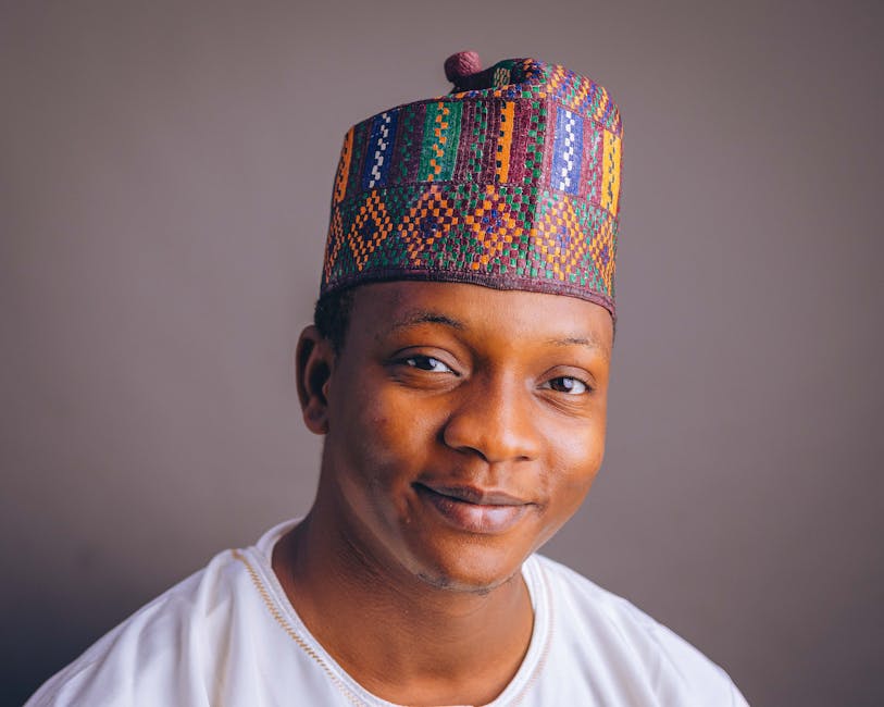 A cheerful young man wearing a colorful Nigerian cap and white attire, smiling softly.
