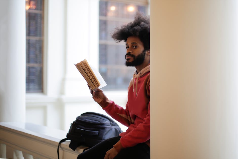 A focused student reading a book while sitting in a university corridor with natural light streaming in.